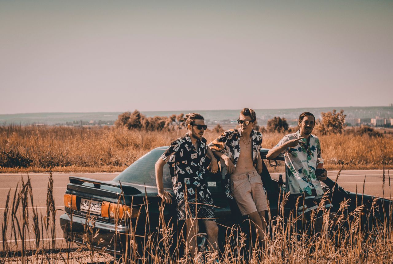 Three young men in printed shirts on a roadside next to a car, enjoying a summer road trip.