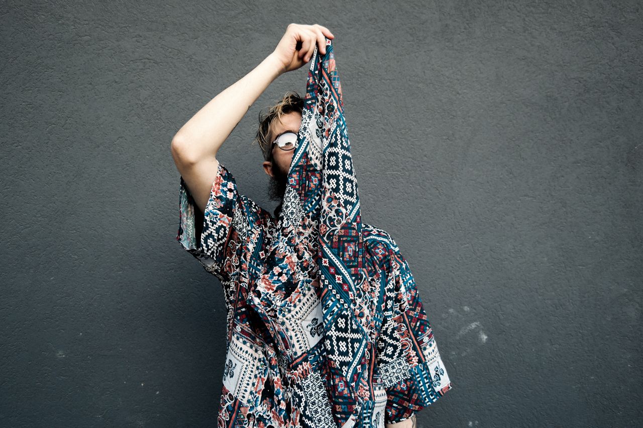 The Art of Drawing Readers In: Your attractive post title goes here A fashionable man with eyeglasses, wearing a printed shirt, uses a bandana to partially cover his face against a plain gray wall.